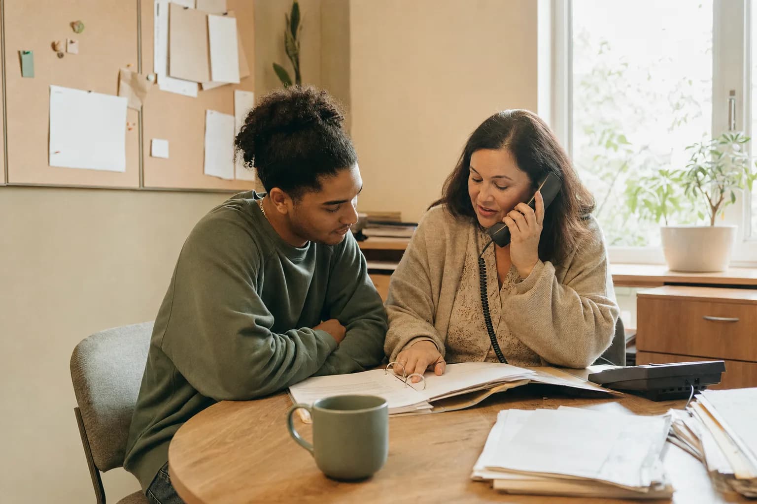 Two adults at a desk with an open folder between them, one making a call